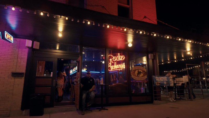 The lit Rodeo Exchange neon sign and front windows on West Exchange Avenue at night, doorman at the entrance